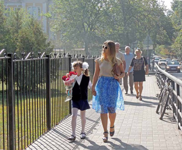 A young adult woman with sunglasses and handbag under arm and a yawning pre-adolescent girl tied in a bows with a bouquet of flowers walk down the sidewalk holding hands