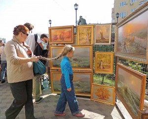 Middle-aged woman holding a plait of elementary age girl trying closer look at the picture on the street exhibition and sale of paintings in the city core during the celebration of the City Day