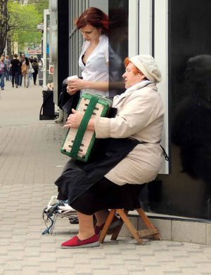 Street mature women musician in the knitted beanie sitting on the wooden camp stool in the city street and playing the accordion. Young women with cup in the hands attentively listens to her standing against the wall
