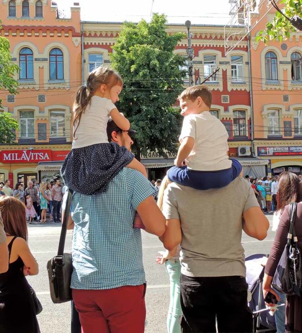 Communication of elementary age girl and boy sitting on fathers shoulders. City dwellers on the parade of Street Theatres on the Platonov Arts Festival